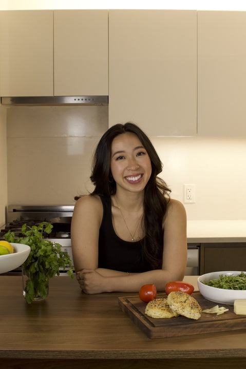 Woman smiling in a kitchen, leaning on a wooden counter with tomatoes, bread, and herbs. Theres a bowl of lemons on the left and a bowl of greens on the right. She is wearing a black sleeveless top.